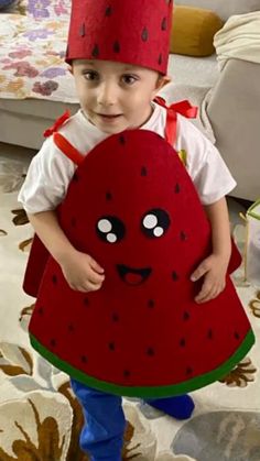 a little boy in a red hat and dress holding a large watermelon pillow