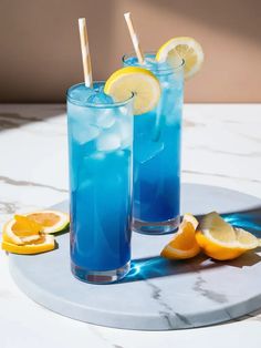 two glasses filled with blue liquid and lemon slices on a marble tray next to sliced oranges