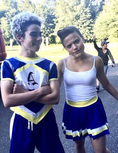 two young people dressed in cheerleader outfits standing next to each other with their arms crossed