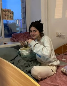 a woman sitting on her bed eating food from a bowl in front of a window