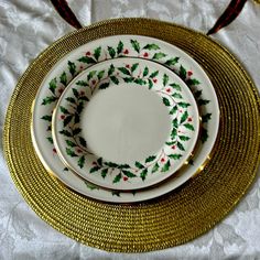 three plates with holly designs on them sitting on a white tablecloth covered table cloth