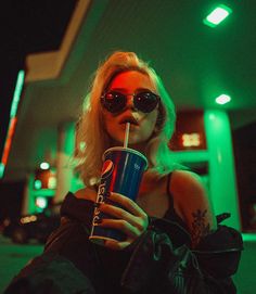 a woman in sunglasses drinking from a blue cup while sitting on the ground at night