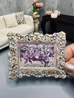 a person holding up a small frame with flowers on it in front of a couch