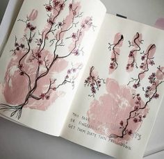 an open book with handprints on it and pink flowers in the middle, sitting on top of a white surface