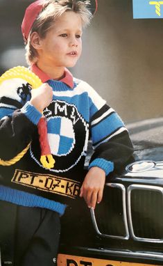 a young boy in a sweater and hat leaning on the hood of a black car