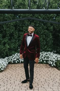 a man in a red velvet jacket and bow tie standing on a brick walkway with white flowers behind him