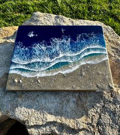 a blue and white tile with waves crashing on the beach in front of grass, rocks and water