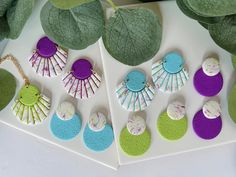 several different colored earrings are displayed on a white surface next to green leafy plants