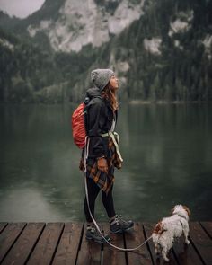 a woman is standing on a dock with her dog and looking up at the mountains