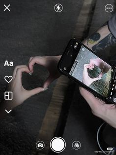 someone is holding their cell phone and making a heart shape with their hands while sitting on the ground