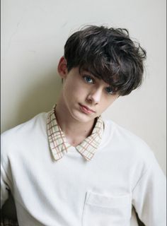 a young man with short hair wearing a white shirt and looking at the camera while standing in front of a wall