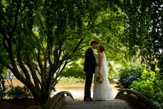 a bride and groom standing on a bridge in the park under some trees with their arms around each other