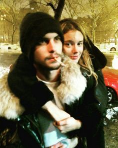 a man and woman standing next to each other in the snow near a red car