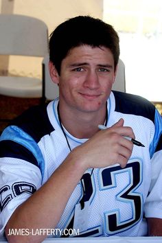 a man sitting at a table with a pen in his hand and wearing a jersey