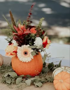 an orange vase with flowers and greenery sits on a table next to pumpkins