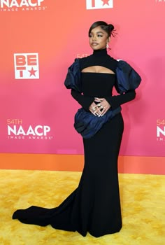 the singer is wearing a black gown with an open neck and long sleeves, while standing on