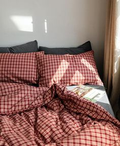 an unmade bed with red and white checkered comforter on it, next to a window