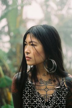 a woman with tattoos and piercings on her chest standing in front of some trees