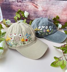 two hats with embroidered flowers on them sitting next to some green leaves and white flowers