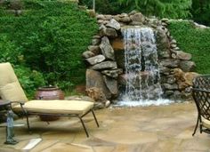 an outdoor patio with chairs, table and waterfall in the backround surrounded by greenery