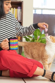 a woman sitting on the floor petting a cat while holding a coffee mug in her hand