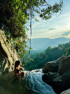 a woman sitting in the middle of a river with rocks and trees around her, surrounded by greenery