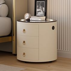 a white round cabinet with two drawers and a clock on the top, next to a bed