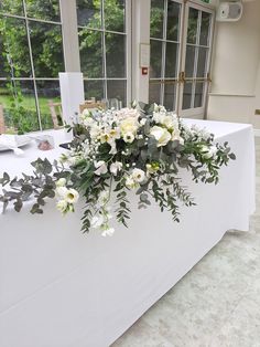 a table with white flowers and greenery on it in front of a large window