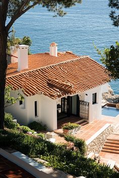 a white house with red tiled roof next to the ocean and trees in front of it