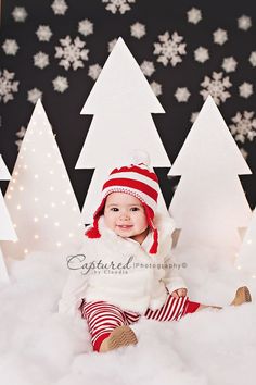 a baby is sitting in the snow surrounded by christmas trees and white lights, smiling at the camera