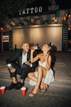 a man and woman sitting on the ground drinking from red cups while one holds up a beer bottle