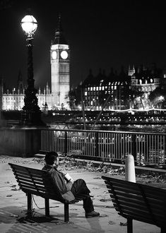 a man sitting on a bench in front of a clock tower at night with the city lights behind him