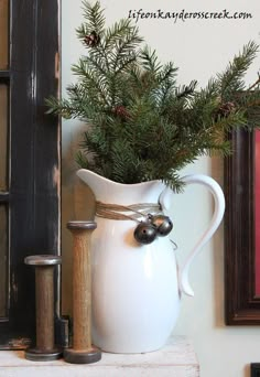 a white pitcher filled with pine branches on top of a mantle