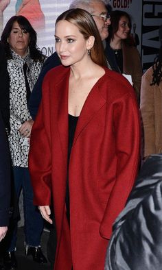 a woman in a red coat and black dress is standing near other people at an event