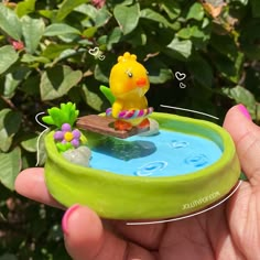 a hand holding a small toy duck in a pond with flowers and plants around it