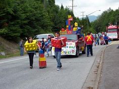 some people are walking down the street with cars and parade floaters in the background