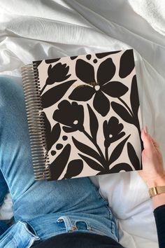 a person is laying on a bed with a notebook in their hand and there is a black and white flower design on the cover