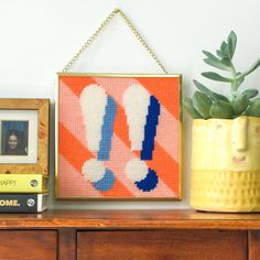 an orange, blue and white cross - stitch pattern hanging on a wall next to a potted plant