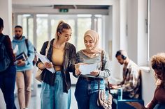 two young women are standing together in an office building, looking at paperwork and smiling