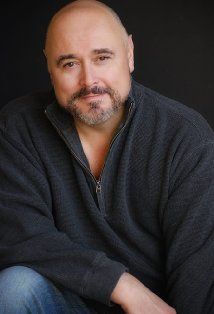 a man with a bald head sitting in front of a black background wearing jeans and a sweater