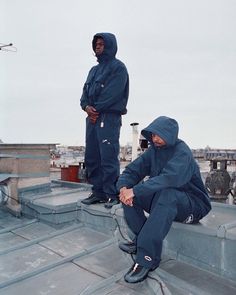 two men sitting on the roof of a building with their feet up and one man standing