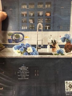 a person holding up a magazine with blue cabinets and flowers in front of them on the counter