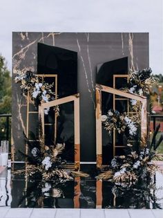 a black and gold wedding backdrop with flowers on the front door, surrounded by greenery