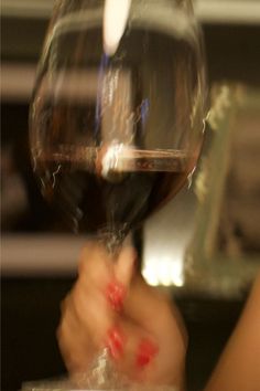 a person holding a wine glass with red liquid in it's hand and blurry background