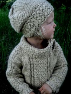 a young child wearing a knitted sweater and hat sitting on the grass in front of some tall grass