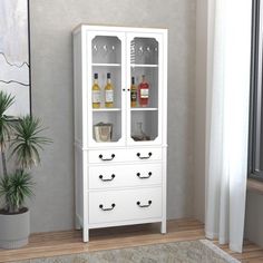 a white cabinet with wine glasses and bottles in it next to a potted plant