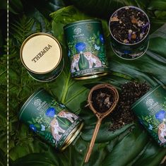 four tins of tea sitting on top of green leaves next to a spoon and cup