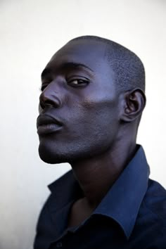 a man with black hair and blue shirt looking off to the side, in front of a white wall