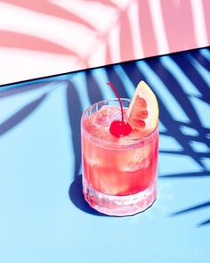 a pink drink with a slice of lemon and a cherry on the rim, sitting on a blue table