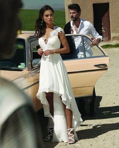 a woman in a white dress standing next to a car with a man looking at her